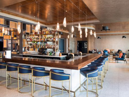 A modern bar interior with sleek blue chairs, pendant lights, and a well-stocked bar, set in a stylish, spacious room.