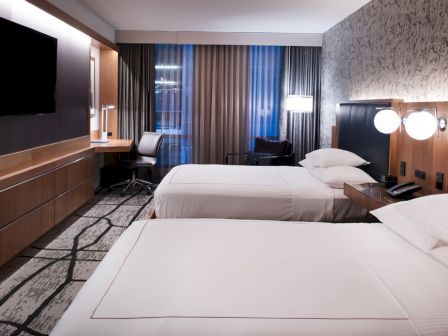 A modern hotel room with two beds, a TV, a desk, lamps, and patterned carpet.