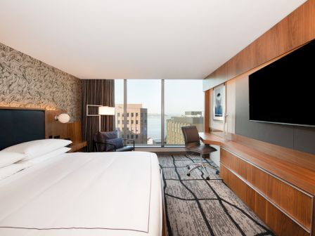 A modern hotel room with a large bed, wall-mounted TV, desk, and a window view of buildings and water in the background.