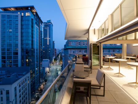A modern balcony overlooks a city street at dusk, with buildings lit up and patio seating visible.