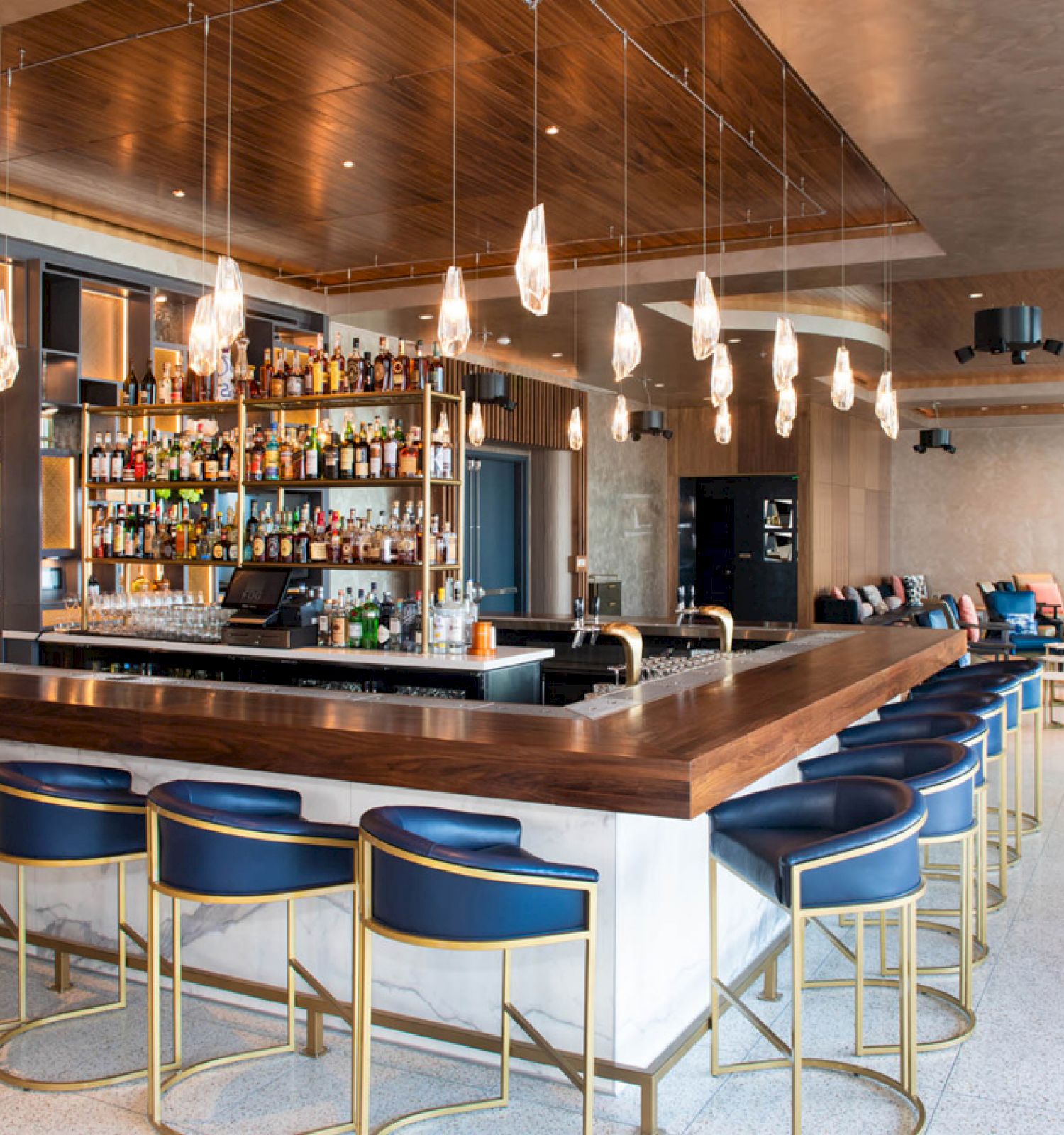 The image shows a modern bar with blue chairs, a wooden countertop, hanging lights, and a fully stocked back wall.