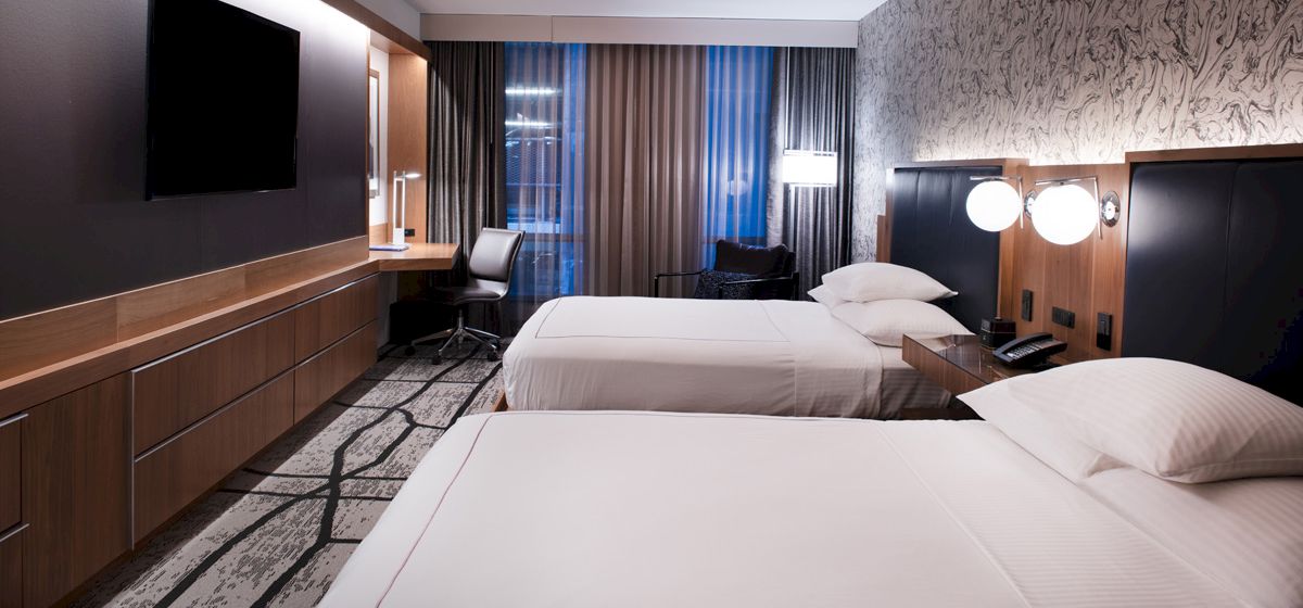 The image shows a modern hotel room with two beds, a flat-screen TV, a desk with a chair, and patterned wallpaper.