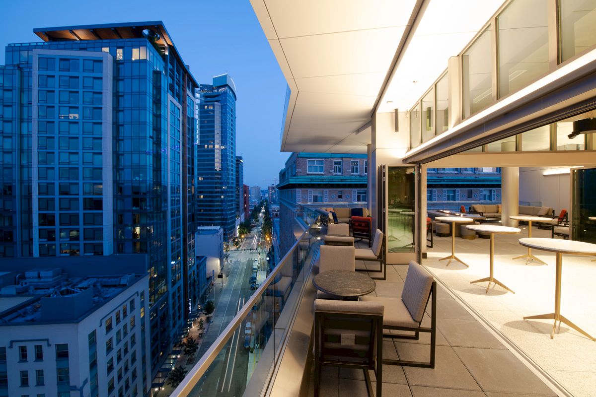 A modern balcony overlooks a cityscape with tall buildings, while tables and chairs are set up for outdoor seating in the evening.