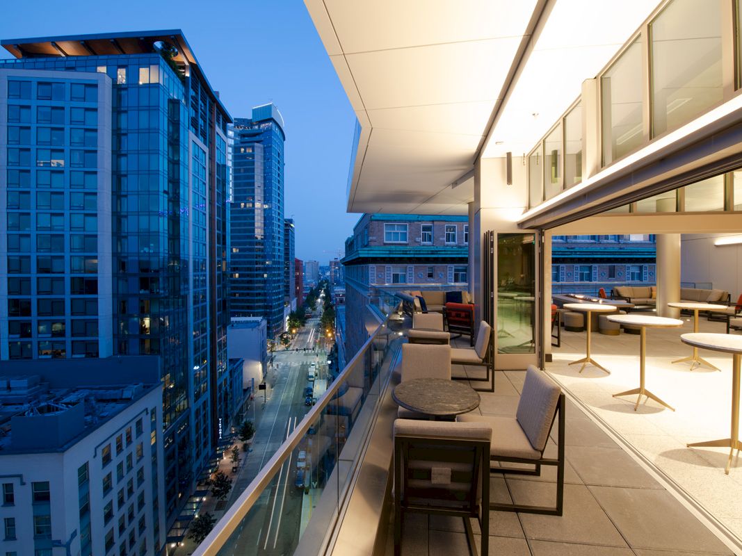A modern balcony overlooks a cityscape with tall buildings, while tables and chairs are set up for outdoor seating in the evening.