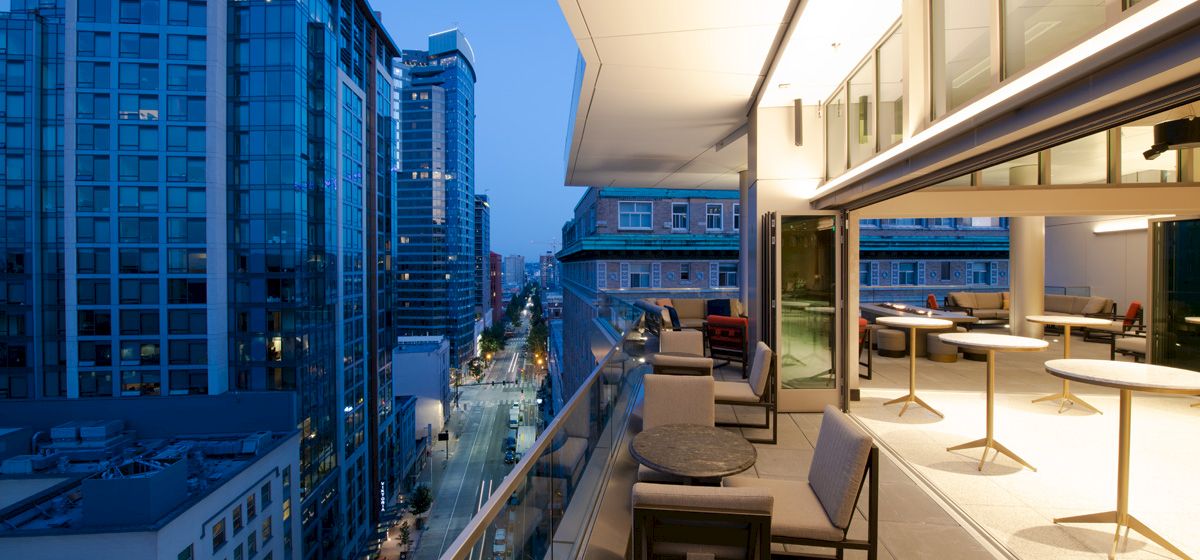 A modern balcony overlooks a cityscape with tall buildings, while tables and chairs are set up for outdoor seating in the evening.