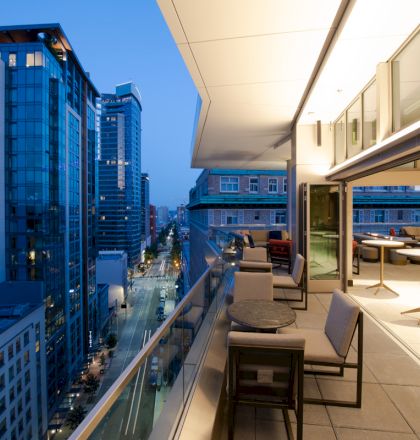 A modern rooftop terrace overlooking a city street with tall buildings. The terrace features seating and tables, lit by ambient lighting.