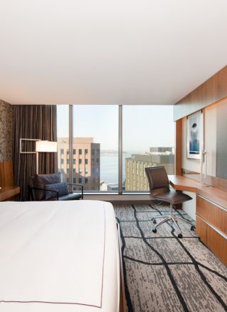 A modern hotel room with a large bed, wall-mounted TV, desk, chair, and window view of city buildings and water.