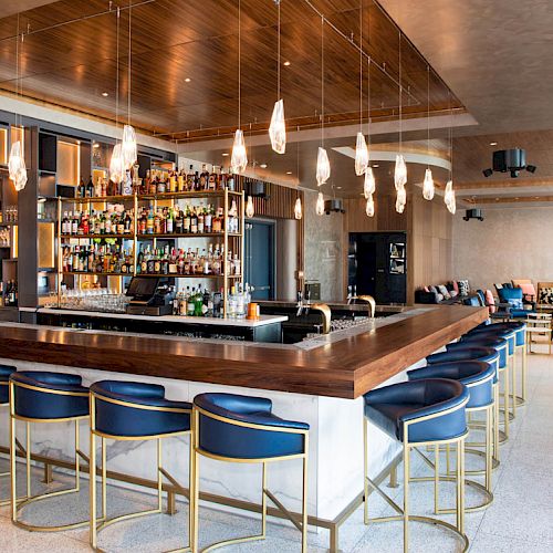 A stylish bar with blue stools, hanging lights, and a fully stocked drink selection. Spacious seating area with modern decor.