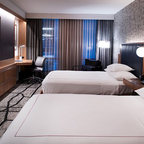 A modern hotel room with two beds, a TV, desk, chair, and stylish decor. The lighting is warm and inviting, and the room looks clean.