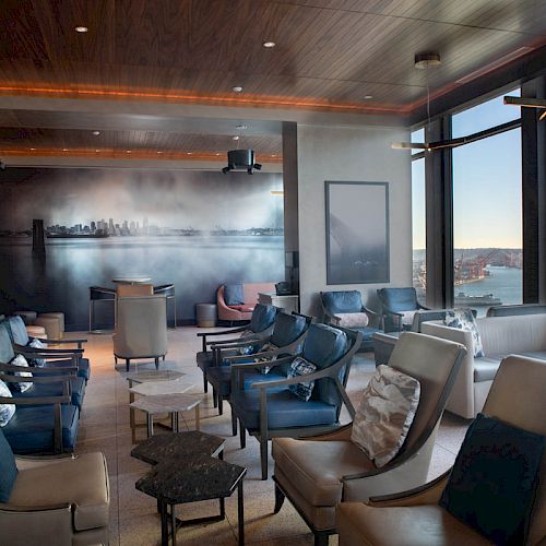 A modern lounge with comfortable seating, a cityscape mural, and large windows offering views of the waterfront and Ferris wheel.