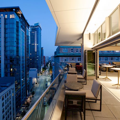 A modern rooftop with seating overlooks a city street lined with tall buildings at dusk. The view includes lit windows and street lights.