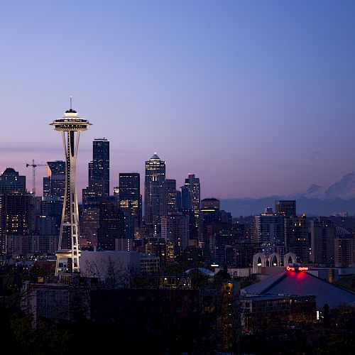 A cityscape of Seattle at dusk, featuring the Space Needle and Mount Rainier in the background, with city lights beginning to illuminate.