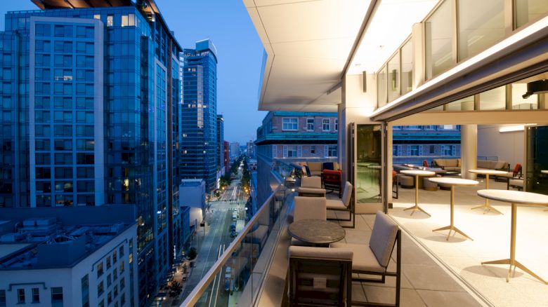 A modern cityscape at dusk with tall buildings and a sleek, stylish balcony featuring seating and tables overlooking the view.