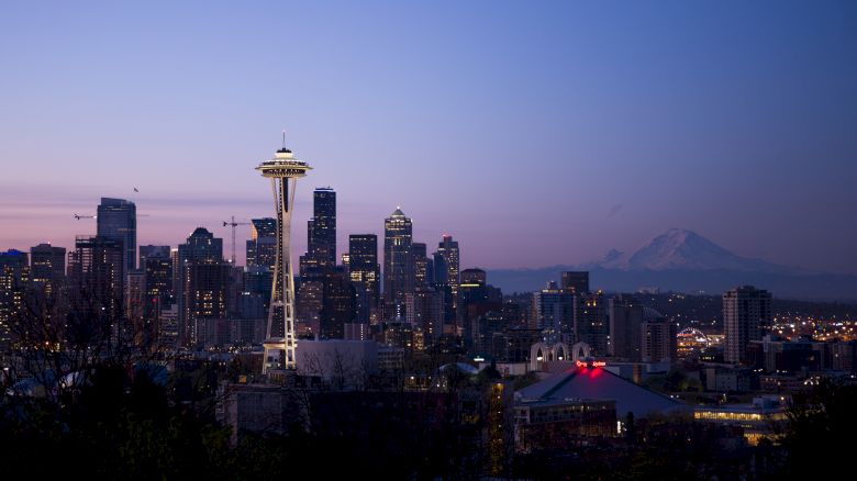 The image shows the Seattle skyline at dusk, featuring the Space Needle and city skyscrapers under a twilight sky with Mount Rainier in the background.