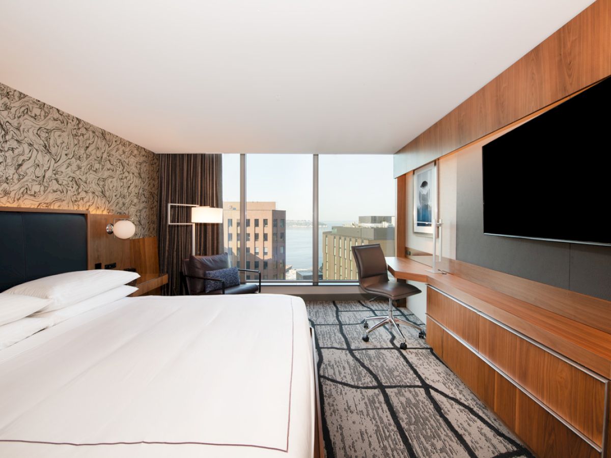A modern hotel room with large bed, desk, chair, and a flat-screen TV. Floor-to-ceiling window offers a cityscape view.