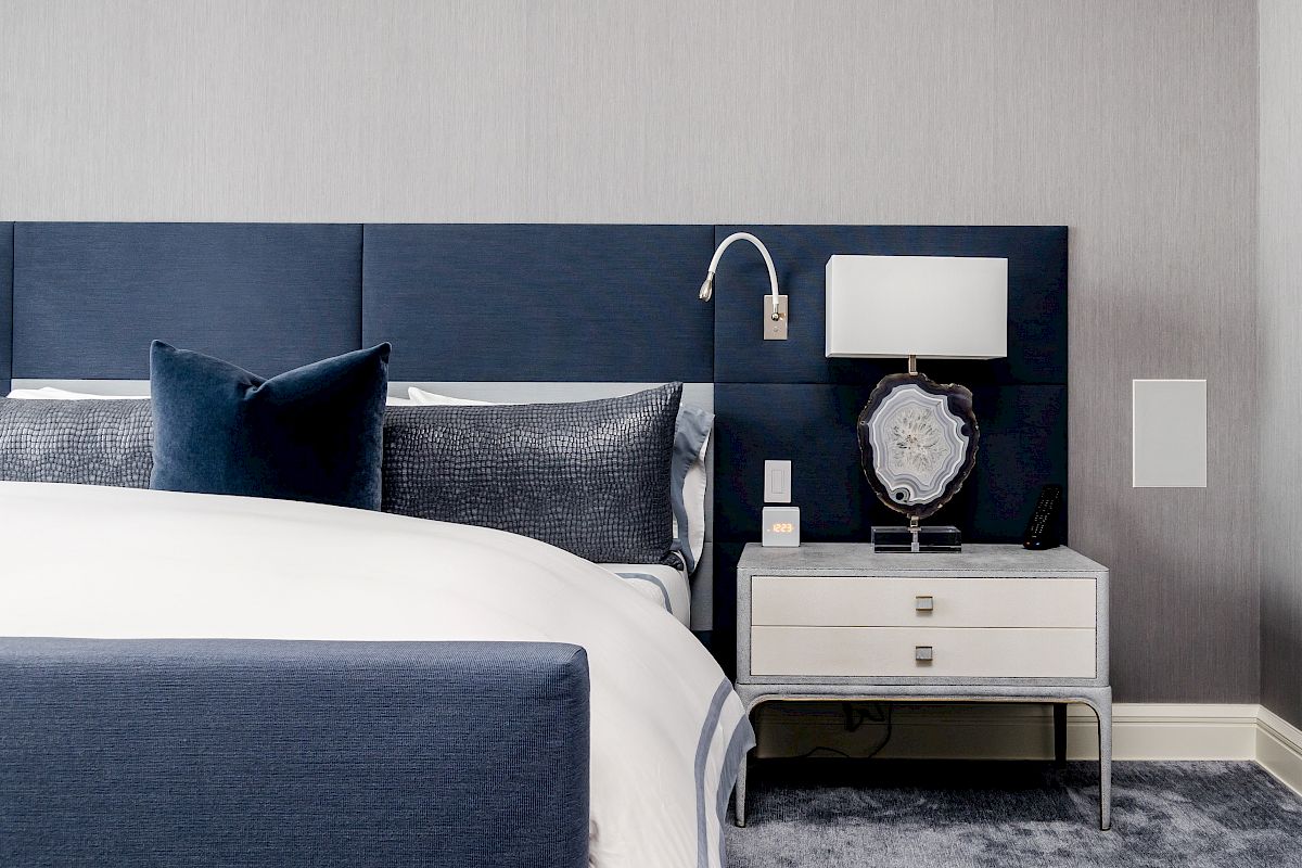 A modern bedroom with a blue headboard, white bedding, and a nightstand featuring a lamp and decorative item on a plush carpet.
