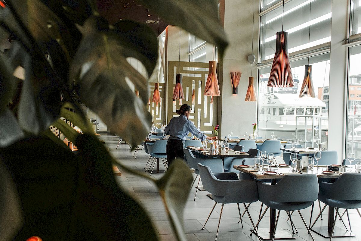The image shows a modern restaurant interior with a person arranging tables, surrounded by plants and upscale decor.