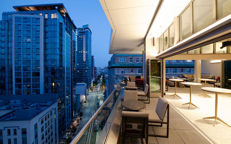 A modern rooftop terrace with seating, overlooking a cityscape of tall buildings at dusk. The scene captures an urban, sophisticated ambiance.