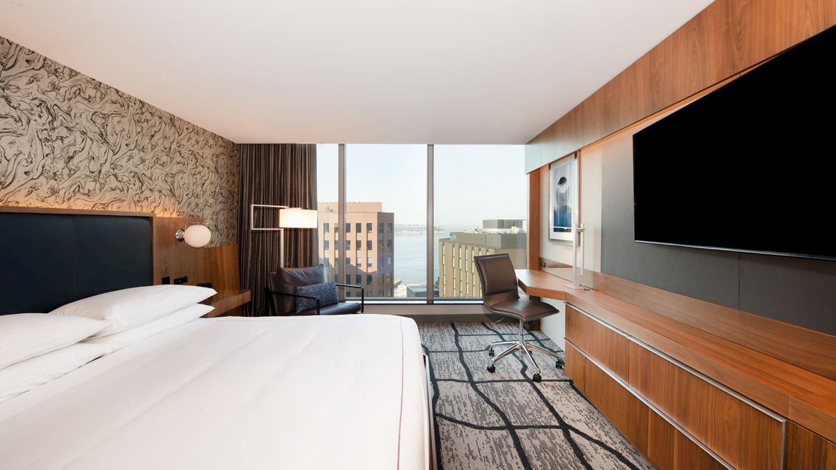 A modern hotel room with a large bed, a desk and chair, and a wall-mounted TV. A window reveals a cityscape and water view.