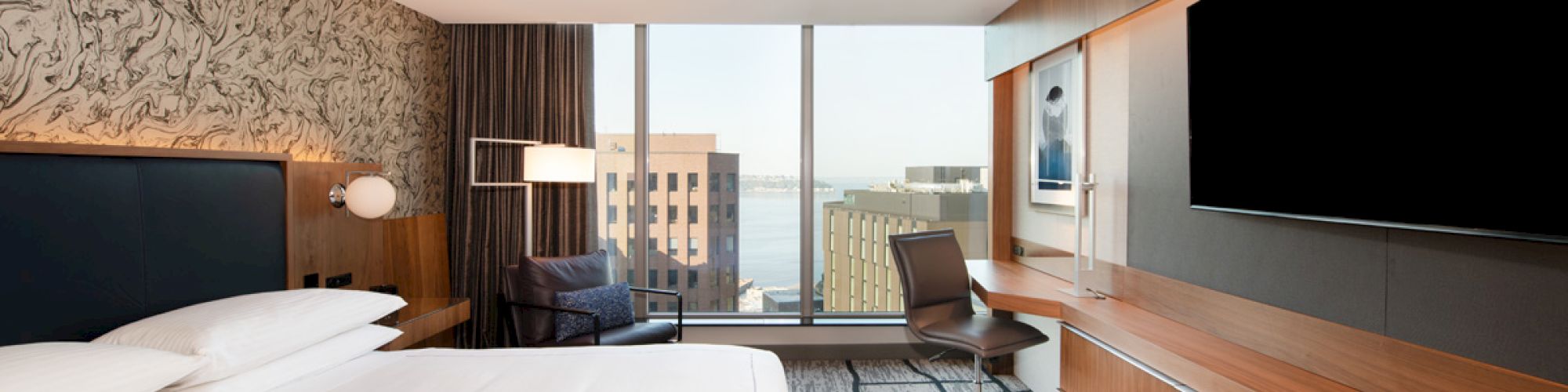 A modern hotel room with a large bed, a desk and chair, and a wall-mounted TV. A window reveals a cityscape and water view.